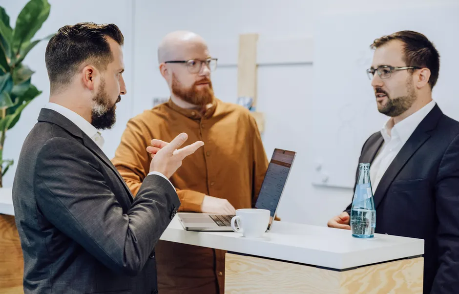 Three men in business attire having a discussion around a standing desk with a laptop and a bottle of water.