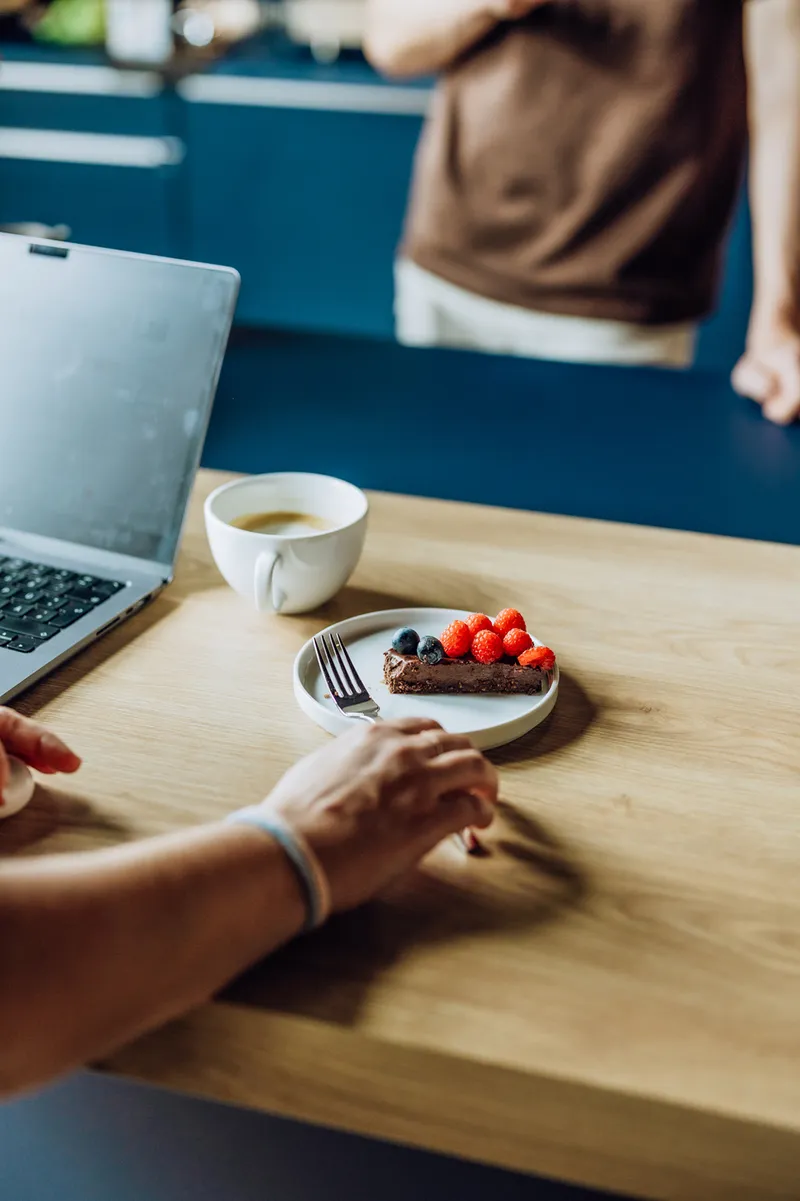 A person reaching for a plate with a slice of chocolate cake topped with berries, next to a cup of coffee and a laptop on a wooden table.