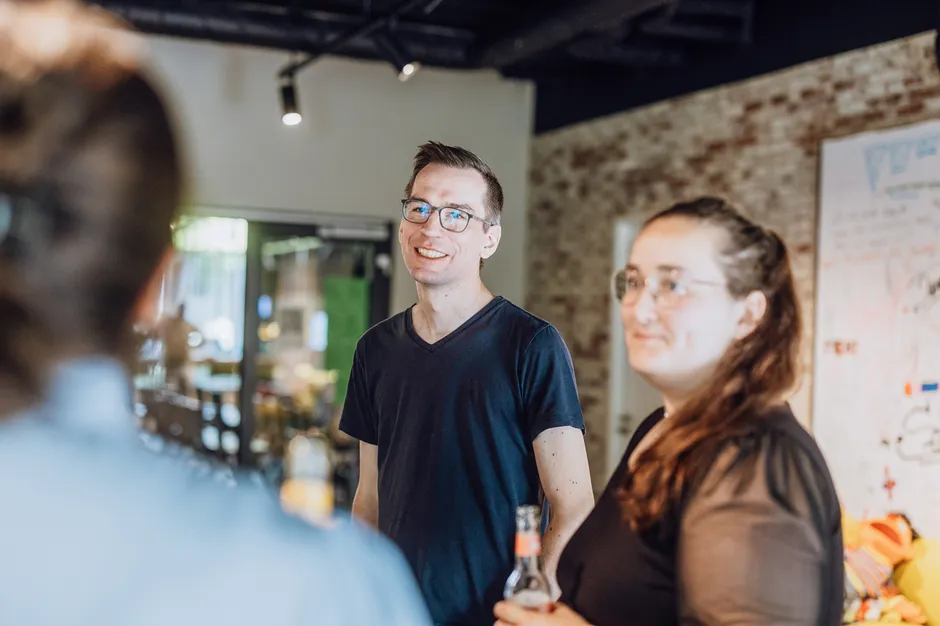 A group of people standing and smiling in a casual indoor setting, holding drinks and engaging in conversation.