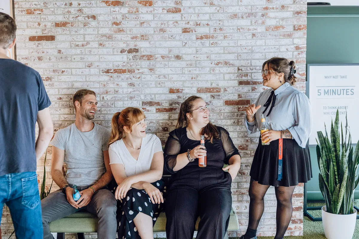 A group of people sitting and standing against a brick wall, smiling and holding drinks, engaged in a casual conversation.