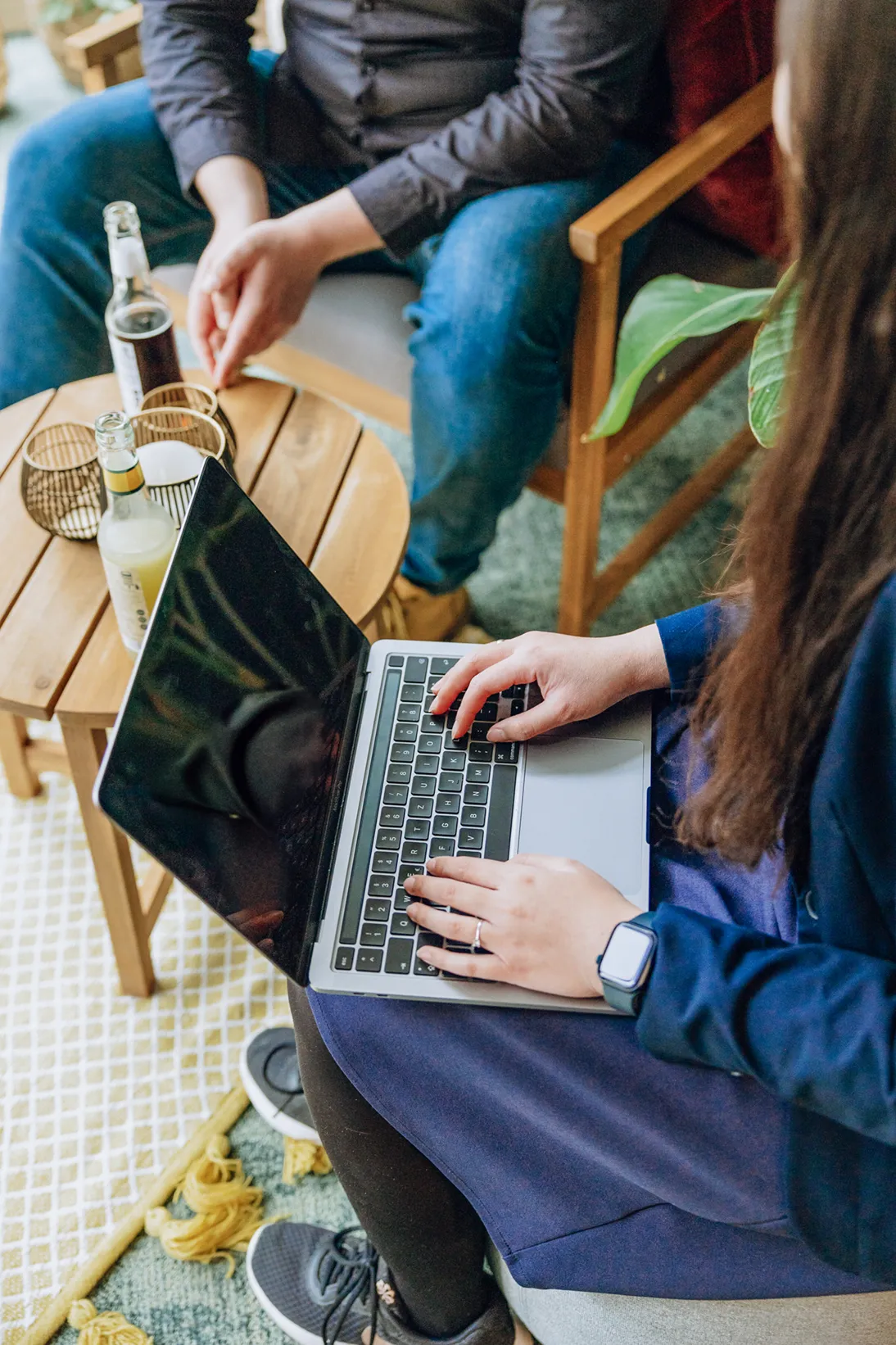Two people sitting at a small wooden table with drinks, one using a laptop and the other with hands clasped.