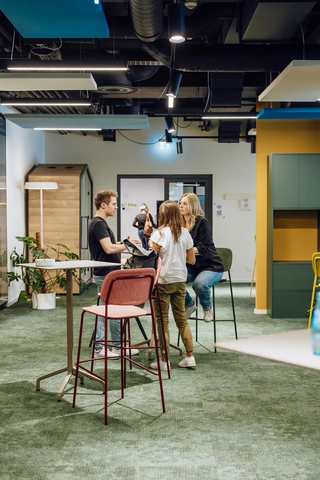 Three people having a casual conversation in a modern office space with high stools and a table.
