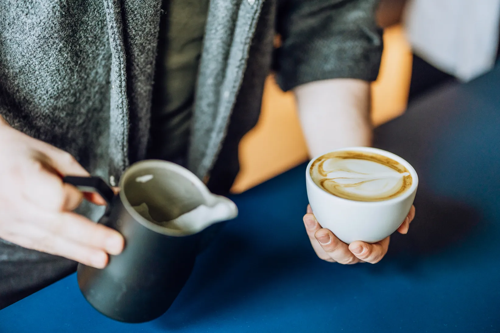 Person holding a cup of coffee with latte art and a milk frothing pitcher on a blue table.
