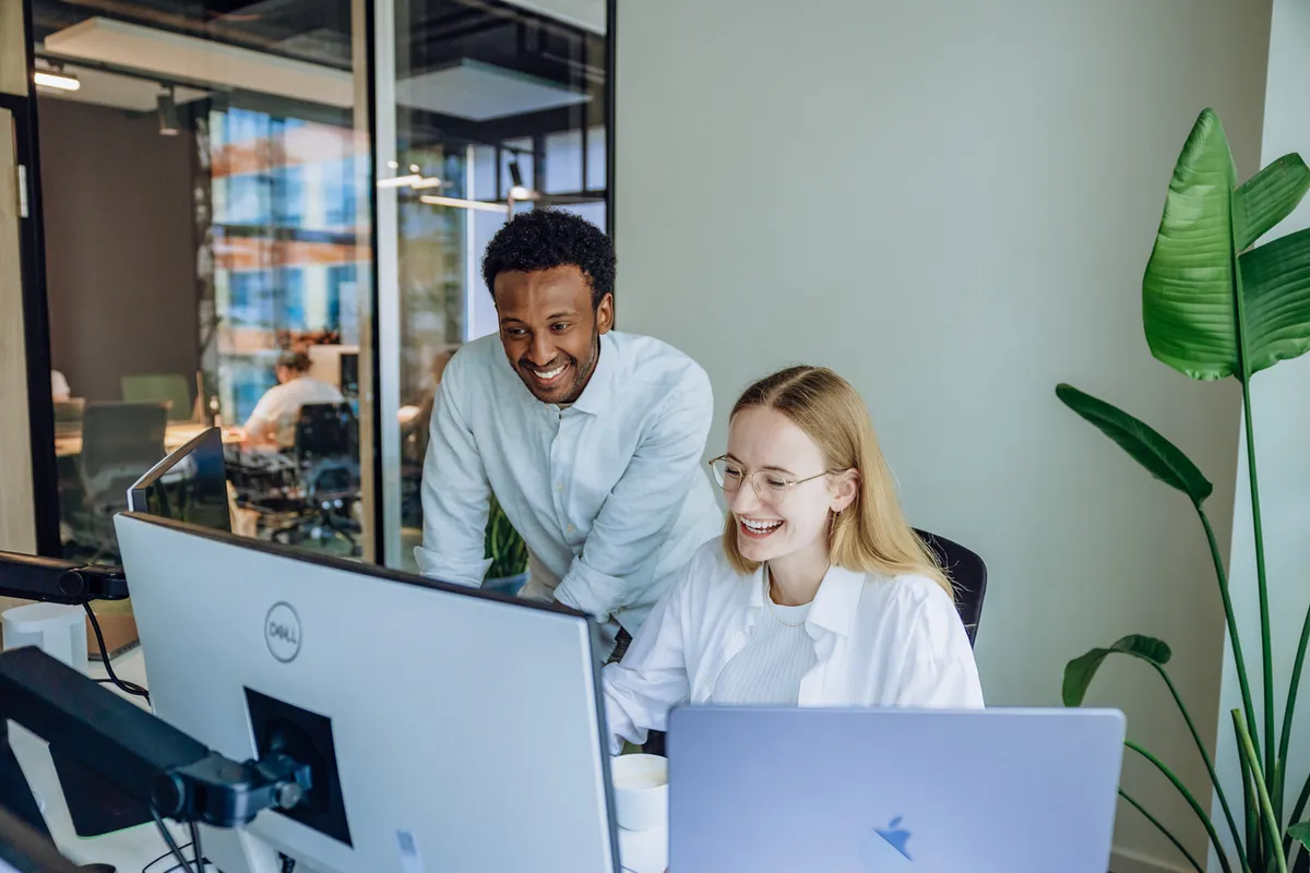 Two colleagues smiling and working together at a desk with computers in a modern office setting.