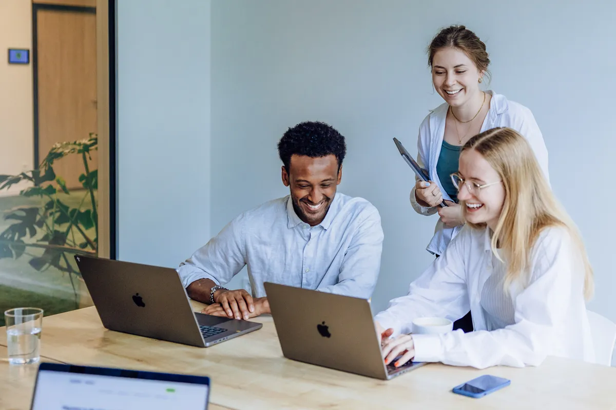 Three colleagues smiling and working together on laptops at a wooden table in a bright office space.