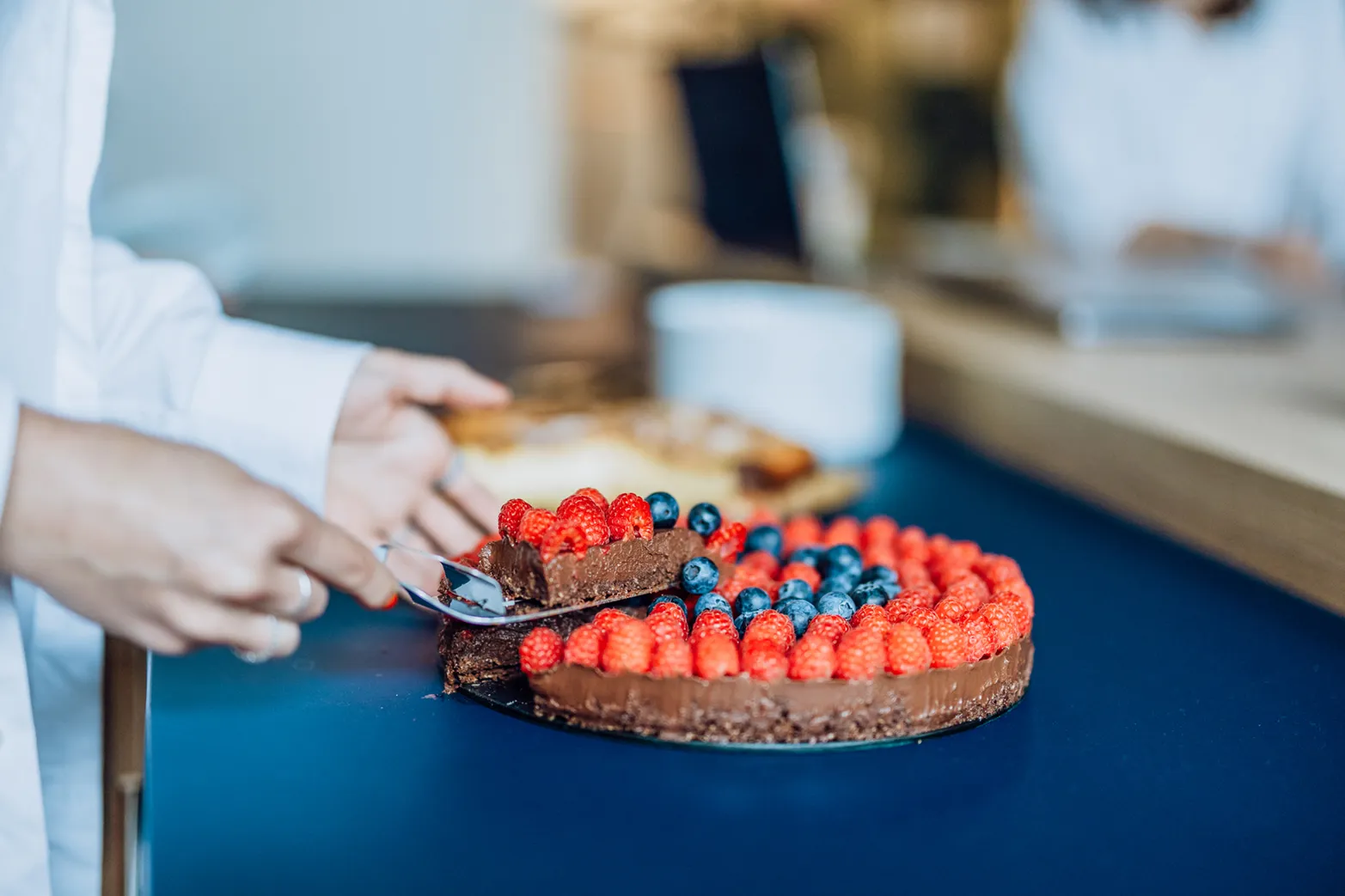 A person cutting a chocolate cake topped with raspberries and blueberries on a blue table.