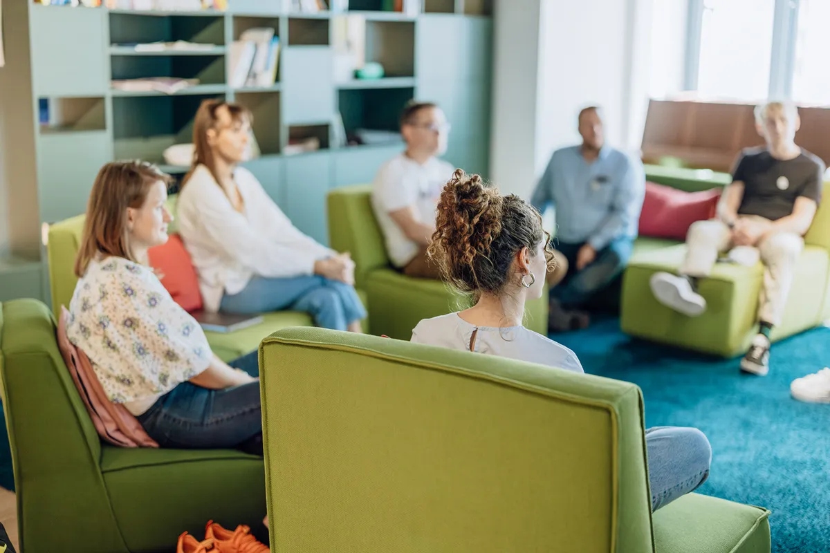 A group of people sitting on green chairs in a cozy office setting, engaged in a discussion. Shelves with books are visible in the background.