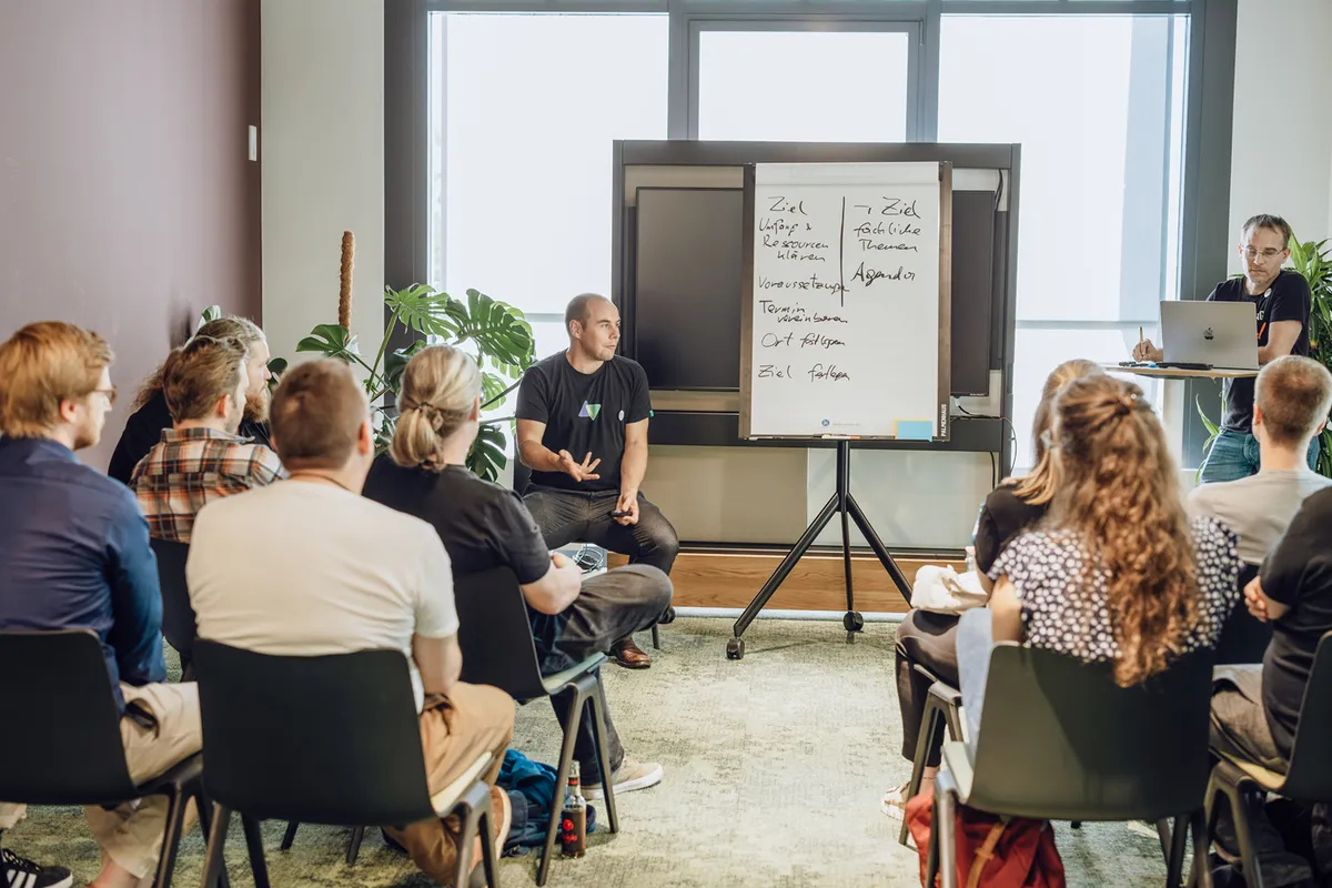 A group of people seated in a room, attentively listening to a speaker who is presenting in front of a flip chart with notes. Another person is standing with a laptop, assisting the presentation.