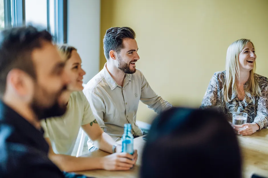 Group of people sitting at a table, smiling and holding drinks, in a casual meeting setting.