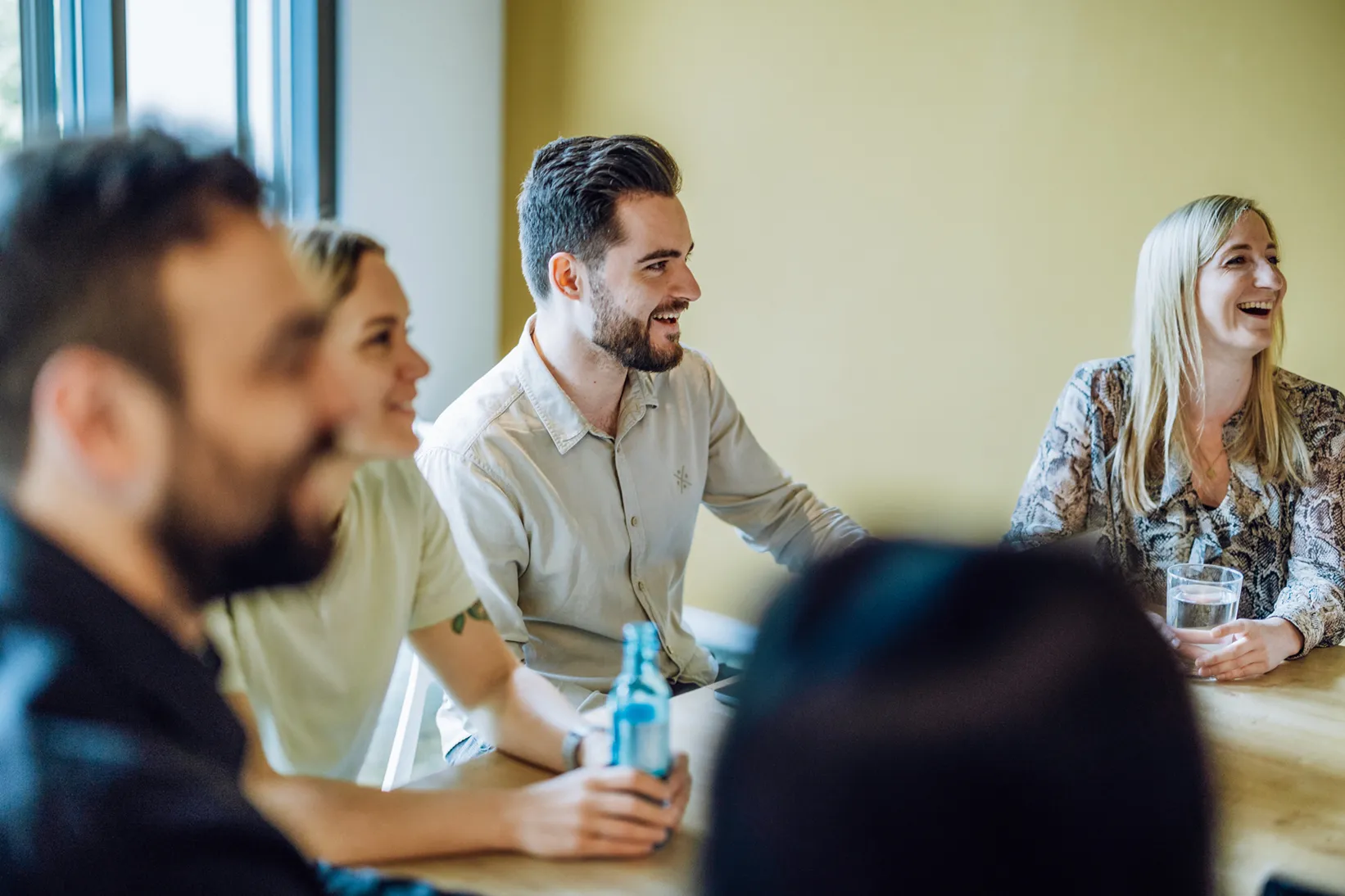 Group of people sitting at a table, smiling and holding drinks, in a casual meeting setting.