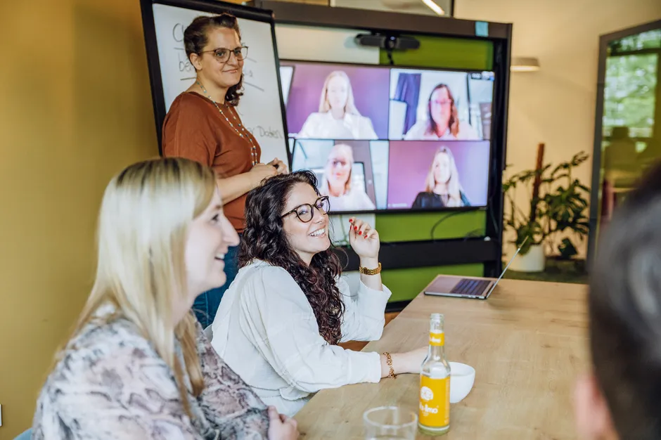 A group of people in a meeting room, with a woman standing near a flipchart and others seated at a table. A video conference screen shows remote participants.