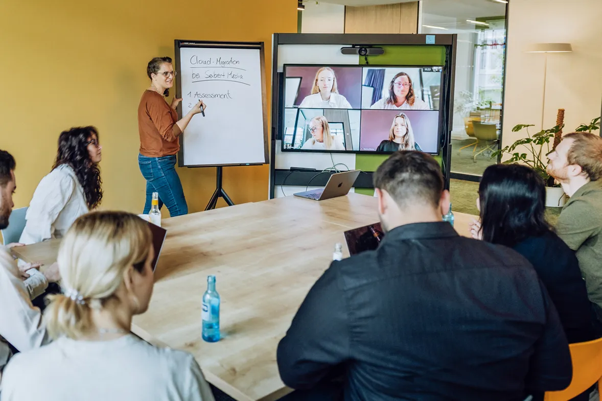 A woman presenting at a flipchart to a group seated around a table, with a video conference screen showing remote participants.