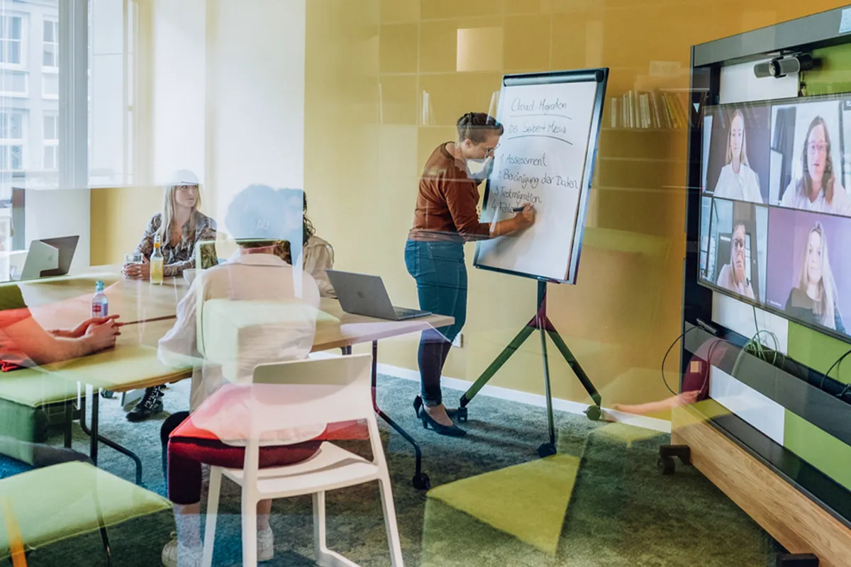 A person writing on a flipchart in a meeting room with several people sitting at a table and a video conference screen showing remote participants.
