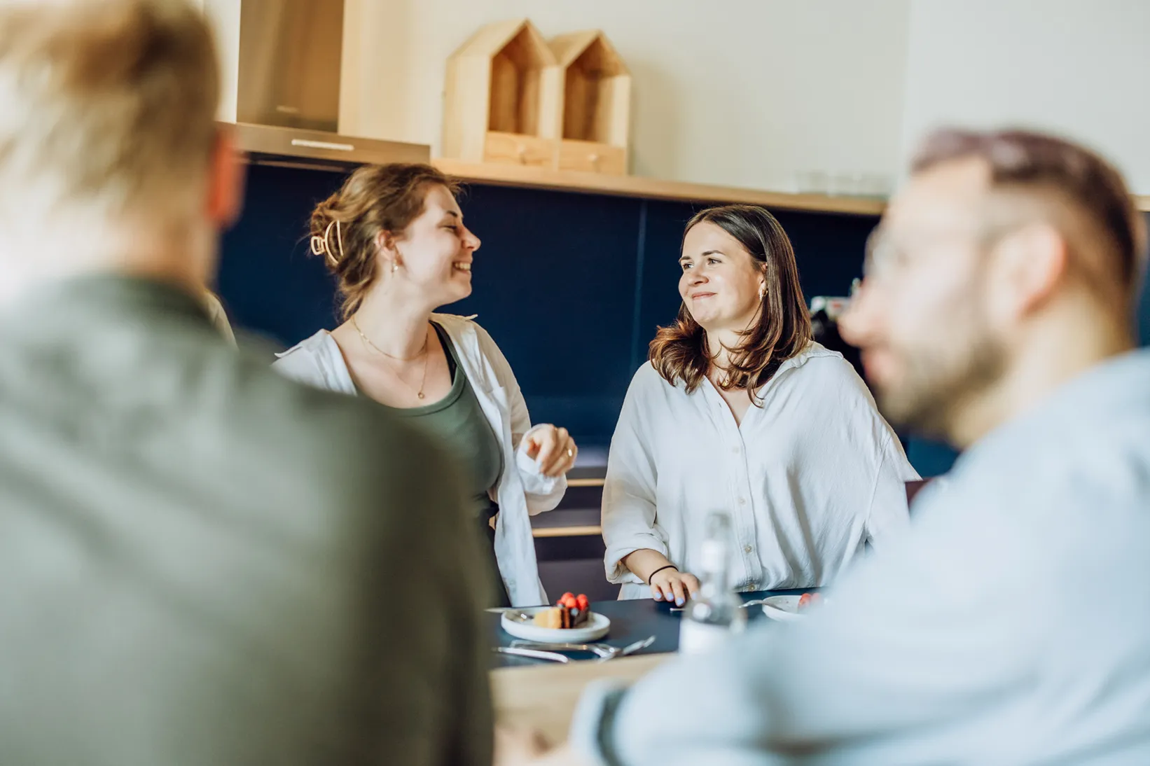 A group of people in a kitchen setting, two women are smiling and conversing while others are blurred in the foreground. A plate with dessert is on the table.