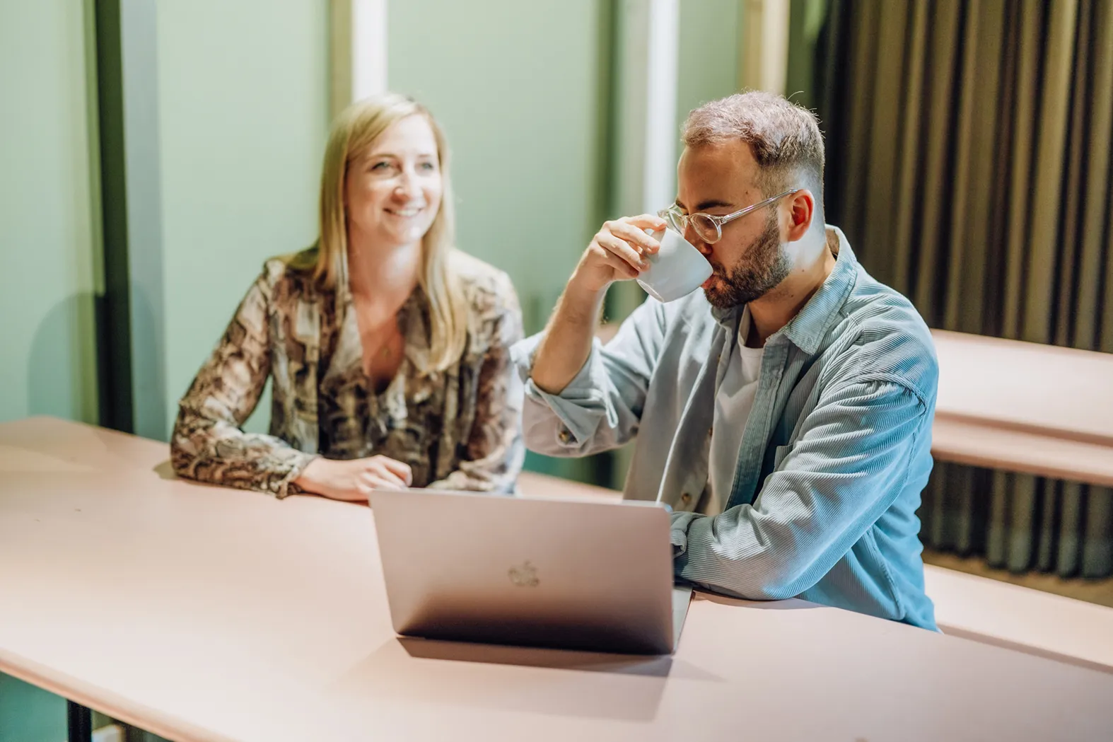A man and woman sitting at a table with a laptop, the man is drinking from a cup while the woman smiles.