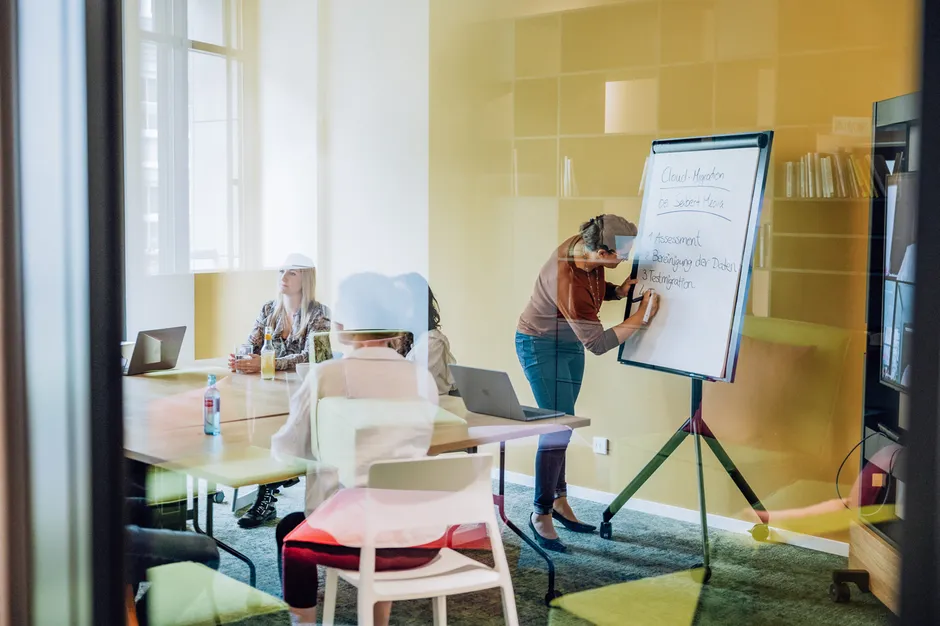 A person writing on a flipchart in a meeting room with several people seated around a table, visible through a glass wall.