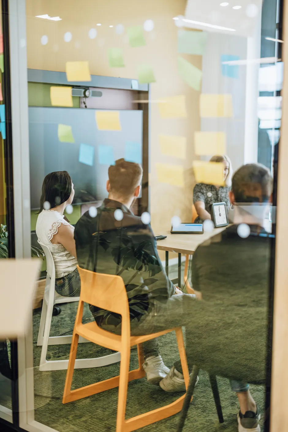 People sitting in a meeting room with colorful sticky notes on the glass wall, viewed from outside.
