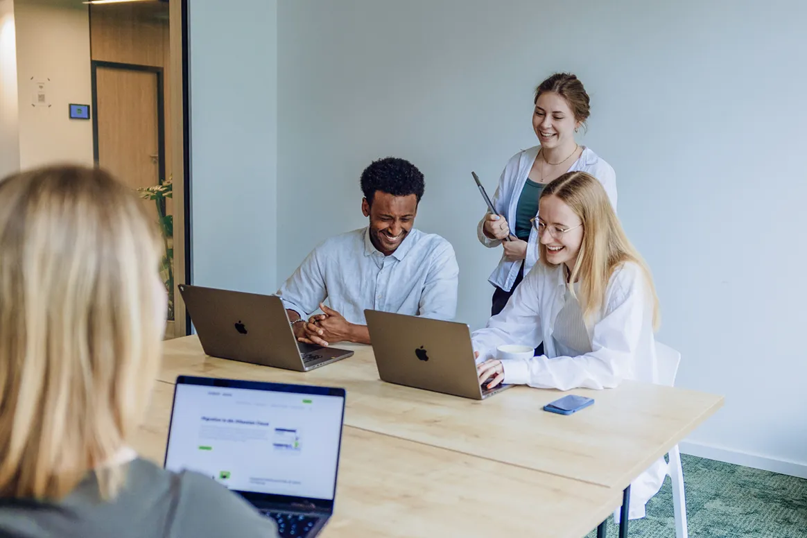A group of four people in an office setting, smiling and working on laptops around a wooden table.