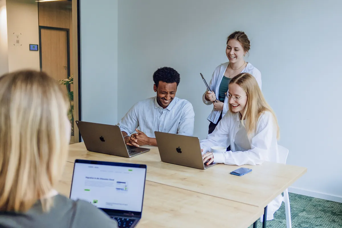 A group of four people in an office setting, smiling and working on laptops around a wooden table.
