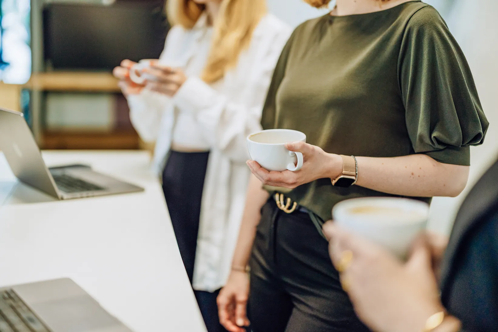 Three people standing with coffee cups near laptops in an office setting.
