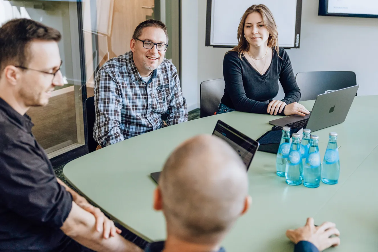 Four people having a meeting around a green table with laptops and bottled water.