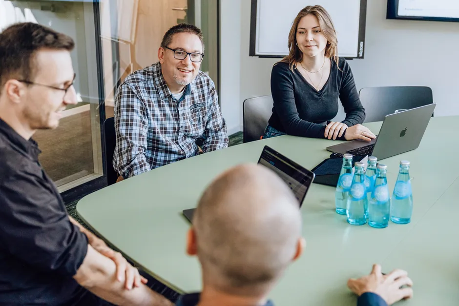 Four people having a meeting around a green table with laptops and bottled water.