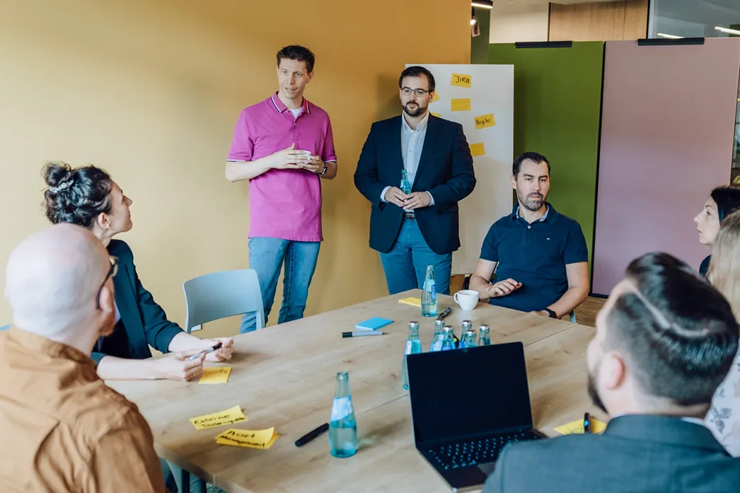 A group of people having a meeting around a table with sticky notes and a laptop. Two men are standing, one in a pink shirt and the other in a suit, while others are seated. A board with sticky notes is visible in the background.