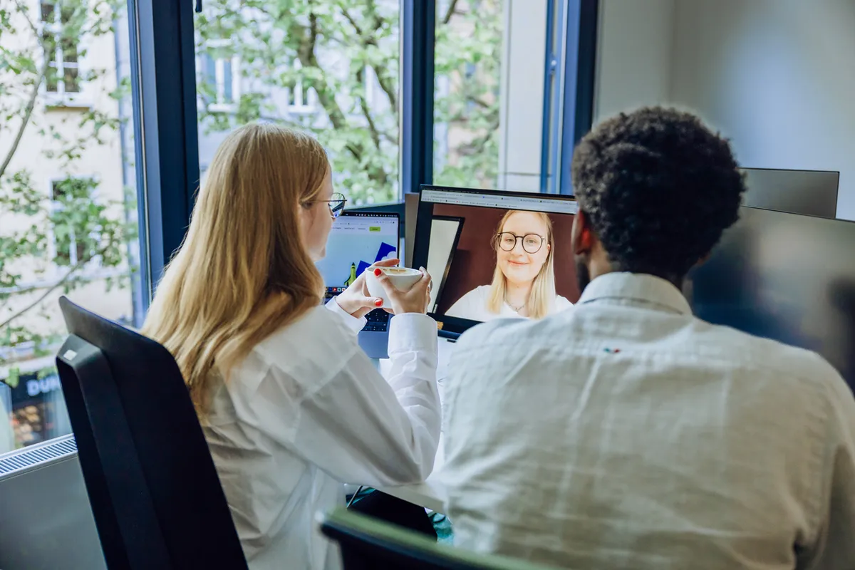 Two people sitting at a desk, engaged in a video call on a computer screen. One person is holding a cup, and the other is looking at the screen. A window with a view of trees is in the background.