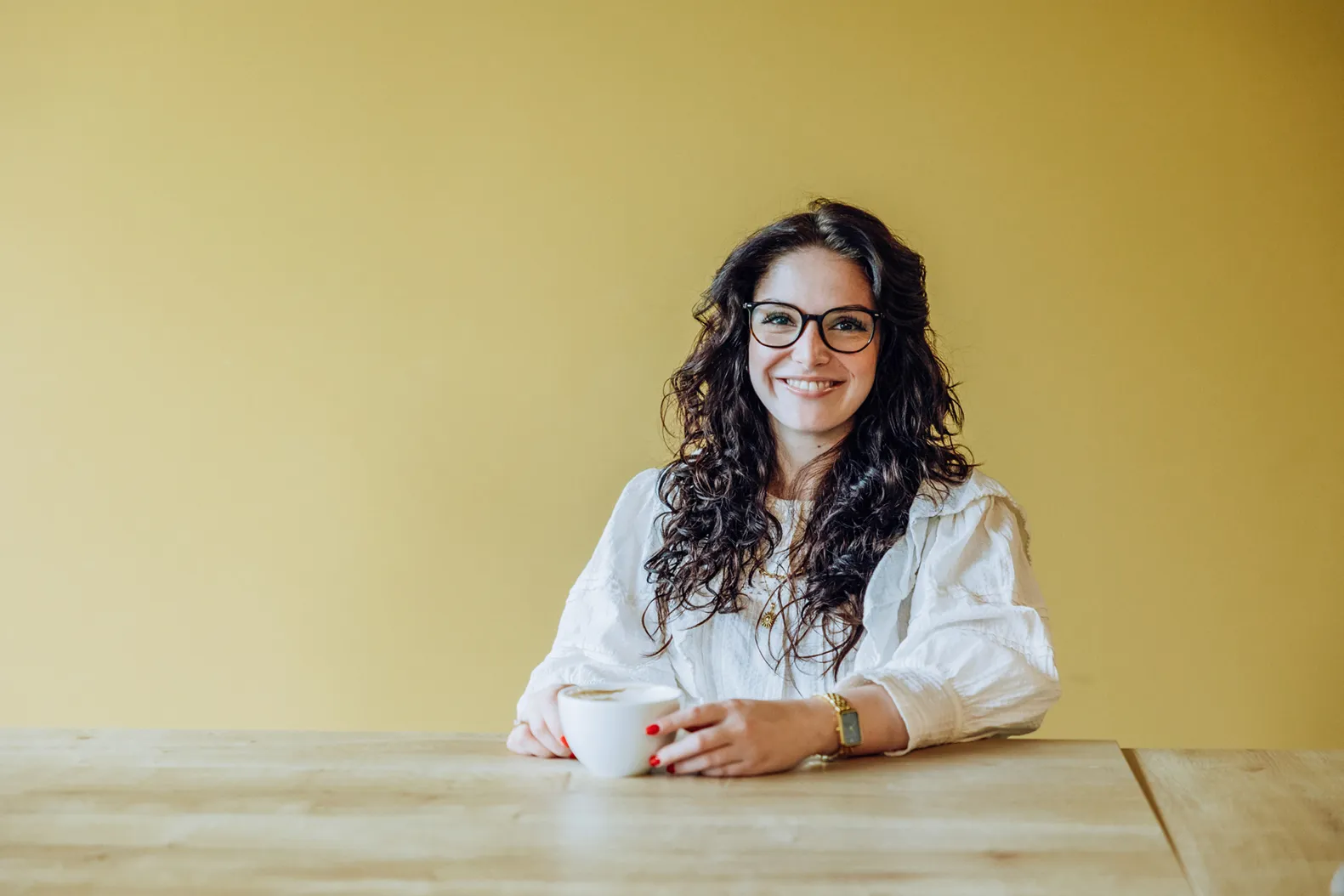 Woman with glasses smiling and holding a coffee cup at a wooden table against a yellow background.