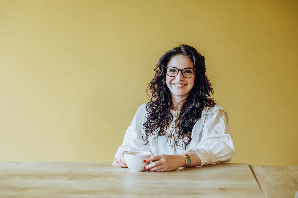 Woman with glasses smiling and holding a coffee cup at a wooden table against a yellow background.
