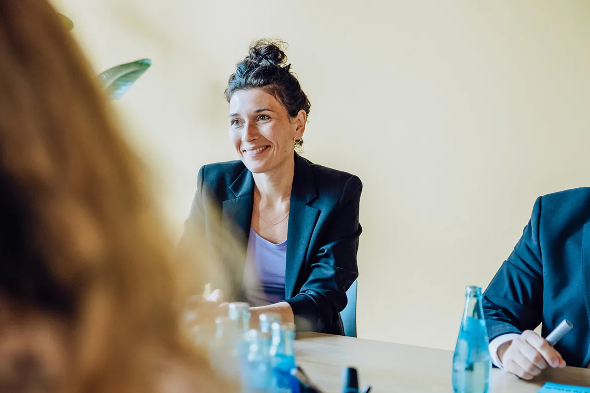 Woman in a blazer smiling during a meeting with water bottles on the table.